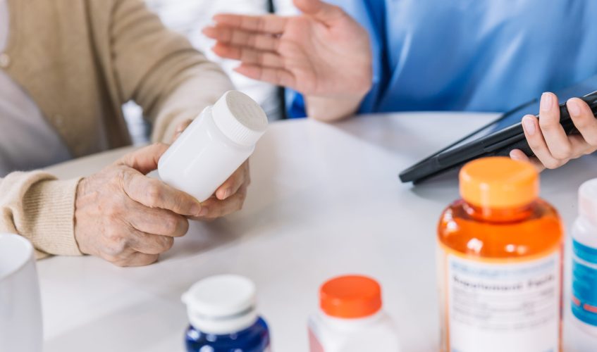 Close up hands of a senior woman holding a medical bottle and asking information from the nurse before administering medication. Caregiver visit at home. Home health care and nursing home concept.
